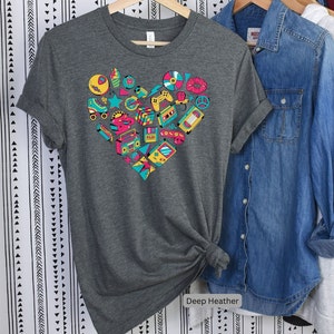 May include: A dark heather grey t-shirt with a heart-shaped design featuring a variety of 80s and 90s icons, including roller skates, cassette tapes, video games, and a boombox. The text "Deep Heather" is printed below the design.