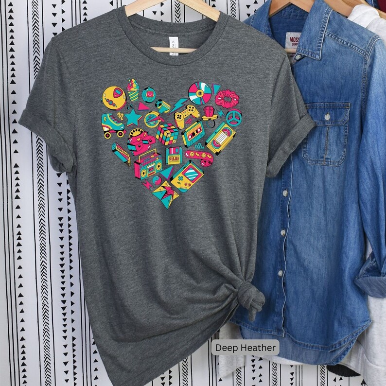 May include: A dark heather grey t-shirt with a heart-shaped design featuring a variety of 80s and 90s icons, including roller skates, cassette tapes, video games, and a boombox. The text "Deep Heather" is printed below the design.