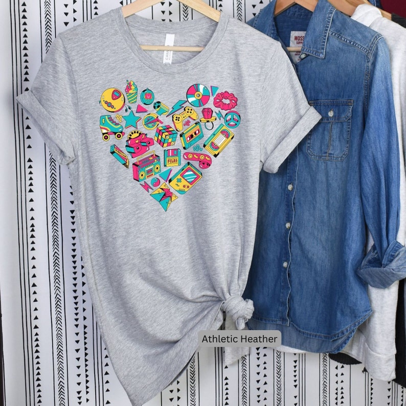 May include: A gray t-shirt with a heart-shaped design made up of various 80s and 90s icons, including a cassette tape, a boombox, a video game controller, and roller skates. The text "Athletic Heather" is printed below the design.