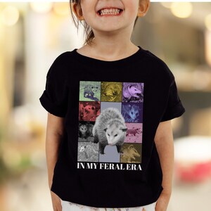 May include: A black t-shirt featuring a colourful graphic of opossums and the text "In My Feral Era".