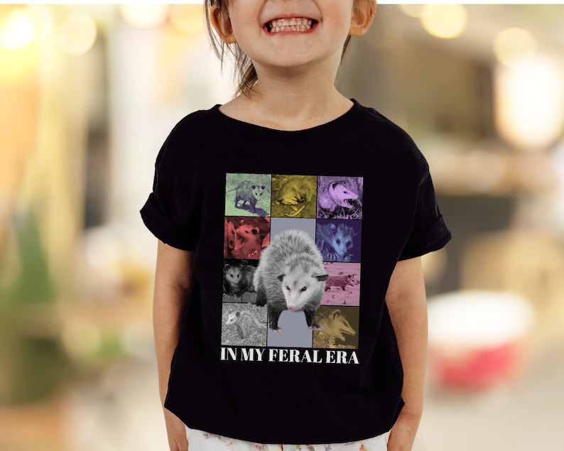 May include: A black t-shirt featuring a colourful graphic of opossums and the text "In My Feral Era".