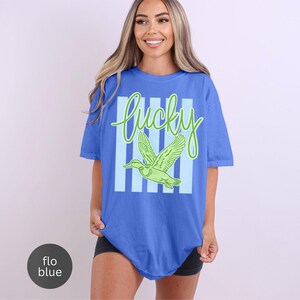 May include: A blue t-shirt with the word "lucky" in green script and a green duck graphic. The word "lucky" is set against a background of light blue vertical stripes. The shirt is a relaxed fit.