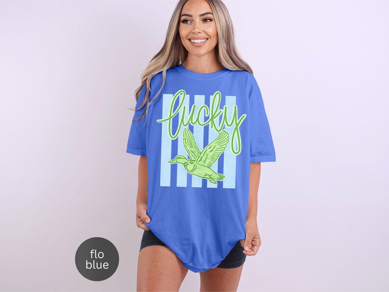 May include: A blue t-shirt with the word "lucky" in green script and a green duck graphic. The word "lucky" is set against a background of light blue vertical stripes. The shirt is a relaxed fit.