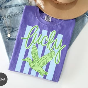 May include: A violet t-shirt with the word "lucky" in green script and a green duck graphic. The shirt is laid on top of blue jeans and a tan hat.