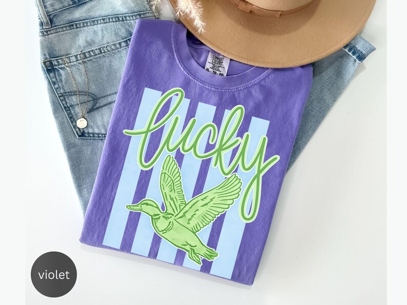 May include: A violet t-shirt with the word "lucky" in green script and a green duck graphic. The shirt is laid on top of blue jeans and a tan hat.