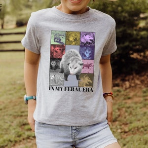 May include: A grey t-shirt featuring a collage of possums in various colours with the text "IN MY FERAL ERA".