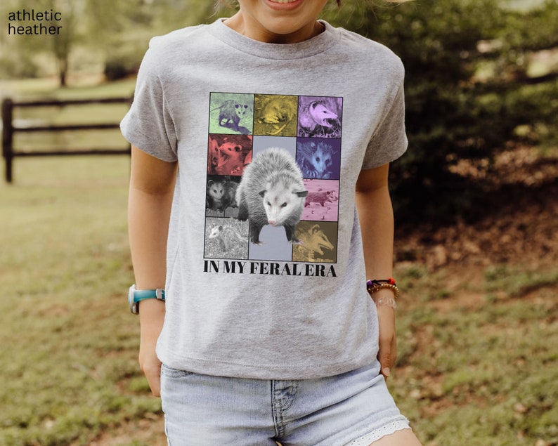 May include: A grey t-shirt featuring a collage of possums in various colours with the text "IN MY FERAL ERA".