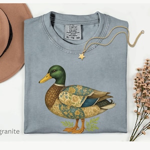 Puede incluir: Una camiseta Comfort Colors azul claro con una ilustraci&oacute;n detallada de un pato. El pato tiene cabeza verde, pico amarillo y alas estampadas. Tambi&eacute;n se ven un collar de estrella dorada y un sombrero marr&oacute;n. La palabra "granite" est&aacute; impresa abajo a la izquierda.