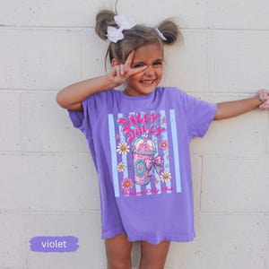 May include: A violet t-shirt with a graphic of a pink drink, daisies, and the text "Dolly Dolly". The shirt has vertical stripes and is worn by a young girl. The word "violet" is written on a small rectangle.
