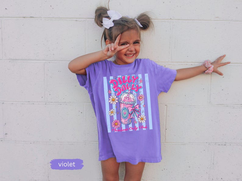 May include: A violet t-shirt with a graphic of a pink drink, daisies, and the text "Dolly Dolly". The shirt has vertical stripes and is worn by a young girl. The word "violet" is written on a small rectangle.