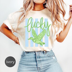 May include: Ivory-colored t-shirt with the word "Lucky" in green script above a green duck graphic. The duck is flying in front of light blue vertical stripes. The shirt is a casual style.