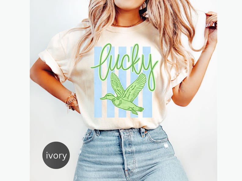 May include: Ivory-colored t-shirt with the word "Lucky" in green script above a green duck graphic. The duck is flying in front of light blue vertical stripes. The shirt is a casual style.