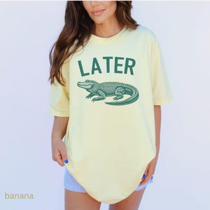 Op de afbeelding: Een geel T-shirt met een groene alligator-print en de tekst "LATER" op de voorkant.