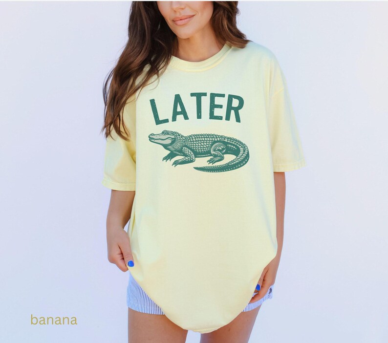 Op de afbeelding: Een geel T-shirt met een groene alligator-print en de tekst "LATER" op de voorkant.
