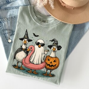 May include: A light green t-shirt featuring a Halloween-themed graphic of three geese dressed in costumes. One goose is a witch, another is a ghost with sunglasses, and the third is a pumpkin. A pink flamingo floatie is also included.