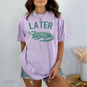 Op de afbeelding: Een lichtpaarse t-shirt met het woord "LATER" bedrukt in groene letters. Onder de tekst staat een groene alligatorafbeelding.