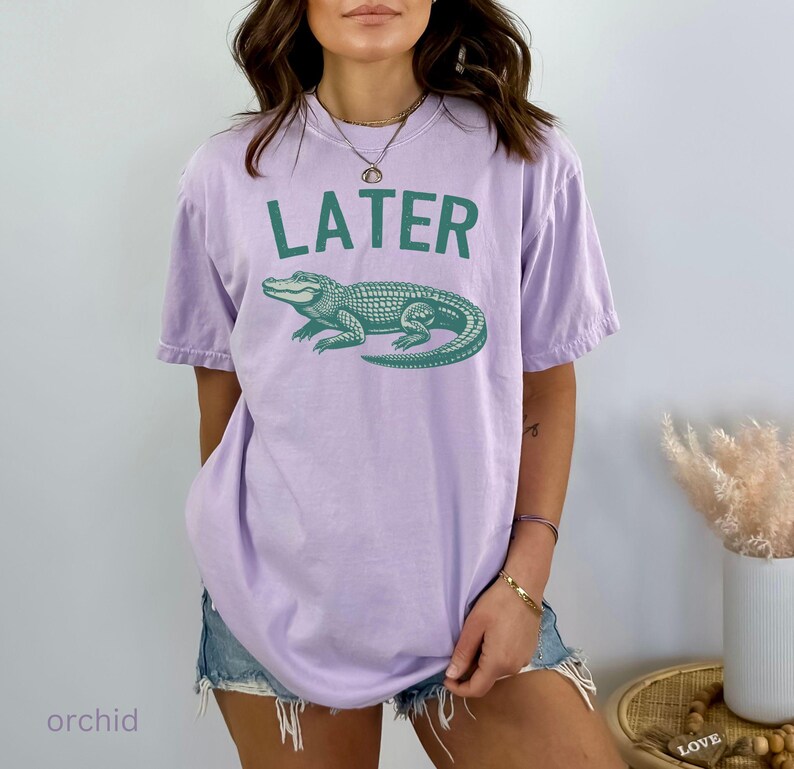 Op de afbeelding: Een lichtpaarse t-shirt met het woord "LATER" bedrukt in groene letters. Onder de tekst staat een groene alligatorafbeelding.