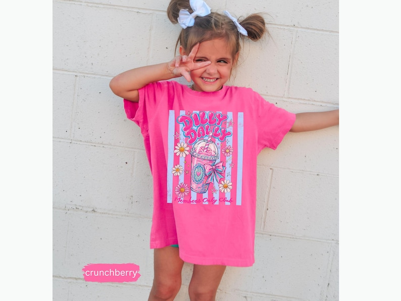 May include: A bright pink t-shirt featuring a graphic of a drink with flowers and the text "Dilly Dally". The shirt is worn by a young person with white bows in their hair.