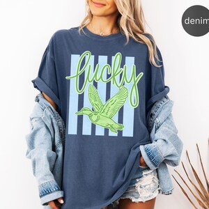 May include: Denim blue t-shirt with a green and light blue graphic. The graphic features the word "Lucky" in a cursive font, a green duck, and light blue vertical stripes. The shirt is part of a casual clothing collection.