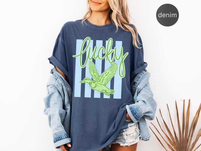 May include: Denim blue t-shirt with a green and light blue graphic. The graphic features the word "Lucky" in a cursive font, a green duck, and light blue vertical stripes. The shirt is part of a casual clothing collection.
