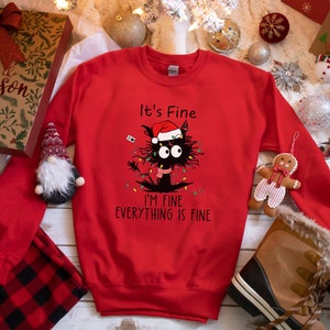 May include: A red sweatshirt with a graphic of a black cat wearing a Santa hat and Christmas lights. Text on the shirt reads "It's Fine, I'm Fine, Everything is Fine." The sweatshirt is surrounded by Christmas decorations.