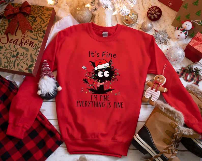May include: A red sweatshirt with a graphic of a black cat wearing a Santa hat and Christmas lights. Text on the shirt reads "It's Fine, I'm Fine, Everything is Fine." The sweatshirt is surrounded by Christmas decorations.