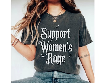 Op de afbeelding: Een donkergrijs T-shirt met de tekst "Support Women's Rage" in wit gedrukt. De tekst is in gotische letter en is omringd door kleine witte sterren.