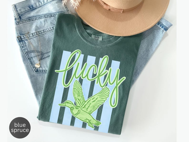 May include: A green t-shirt with the word "lucky" in a light green script, and a green duck graphic. The shirt is laid on top of blue jeans and a tan hat. A gold necklace is visible.