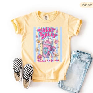 May include: Yellow t-shirt with a graphic of a pink drink, daisies, and the text "Dilly Dally Club Members Only Club." The shirt is paired with denim shorts and black and white checkered slip-on shoes.