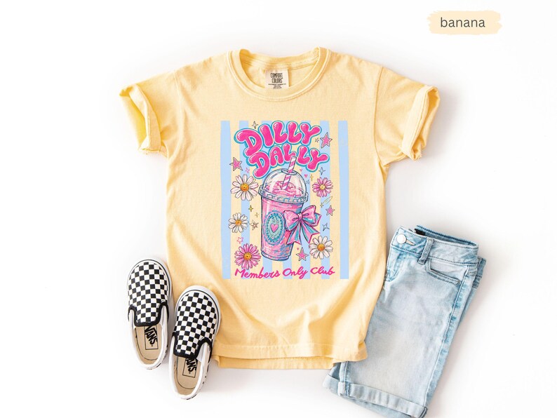 May include: Yellow t-shirt with a graphic of a pink drink, daisies, and the text "Dilly Dally Club Members Only Club." The shirt is paired with denim shorts and black and white checkered slip-on shoes.