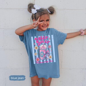May include: A blue jean t-shirt with a graphic of a pink drink with the text "Dilly Dally". The shirt has vertical white stripes and floral accents. The shirt is worn by a young person.