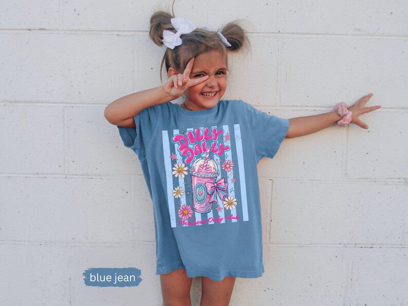 May include: A blue jean t-shirt with a graphic of a pink drink with the text "Dilly Dally". The shirt has vertical white stripes and floral accents. The shirt is worn by a young person.
