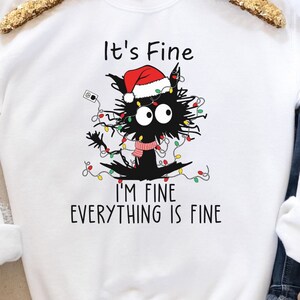 May include: White sweatshirt featuring a black cat tangled in Christmas lights, wearing a Santa hat and scarf. Text reads "It's Fine, I'm Fine, Everything is Fine". A humorous holiday-themed graphic.