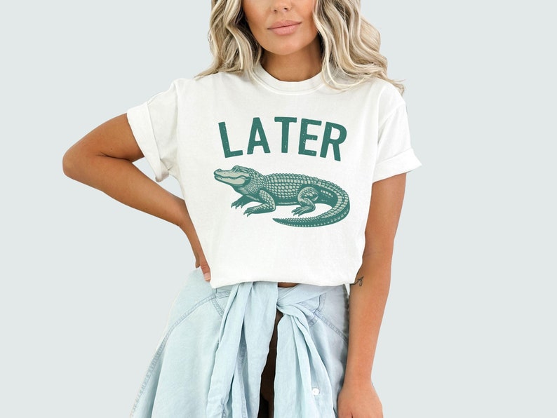 Op de afbeelding: Wit T-shirt met een groene alligator-afbeelding en het woord "LATER" in het groen.