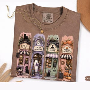 May include: An espresso-colored t-shirt featuring a graphic print of four storefronts: Black Cat Café, Ghost Bookstore, Witch Bakery, and Pumpkin Florist. The shirt has a Comfort Colors label at the neck and is displayed with a gold star necklace.