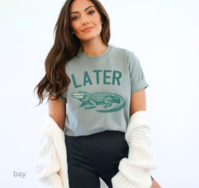 Op de afbeelding: Een lichtgrijs T-shirt met een groene alligator-afbeelding en de tekst "LATER" gedrukt op de voorkant.