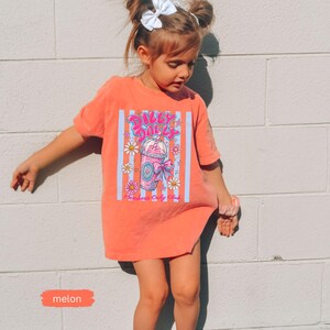 May include: A melon-colored t-shirt with a graphic of a pink drink, daisies, and the text "Silly Daily." The shirt has a vertical striped design in blue and white. The word "melon" is written on a small orange rectangle.