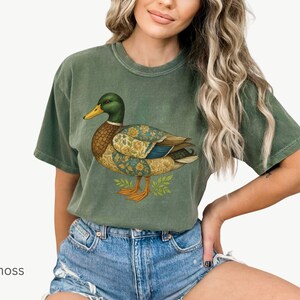 Puede incluir: Camiseta verde musgo con una ilustraci&oacute;n de un pato. El pato tiene cabeza verde, pico amarillo y cuerpo con patrones florales dorados y azules. La camiseta es de corte holgado.