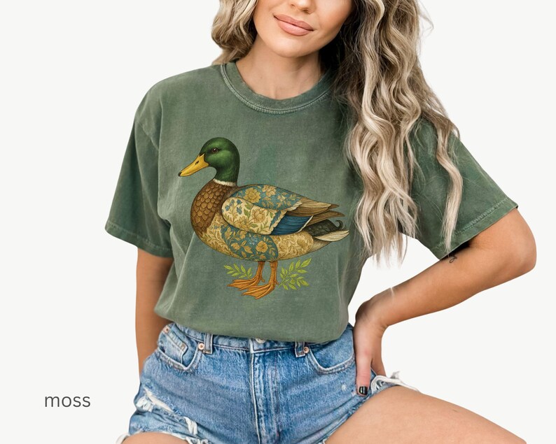 Puede incluir: Camiseta verde musgo con una ilustraci&oacute;n de un pato. El pato tiene cabeza verde, pico amarillo y cuerpo con patrones florales dorados y azules. La camiseta es de corte holgado.