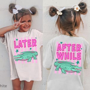 Puede incluir: Camiseta blanca con un dise&ntilde;o de cocodrilo rosa y verde. La parte delantera de la camiseta dice "LATER" en rosa, y la parte trasera dice "AFTER WHILE". La camiseta la lleva un ni&ntilde;o peque&ntilde;o.