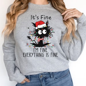 May include: A light gray sweatshirt featuring a black cat tangled in Christmas lights and wearing a Santa hat and scarf. The text "It's Fine, I'm Fine, Everything is Fine" is printed above and below the cat illustration.