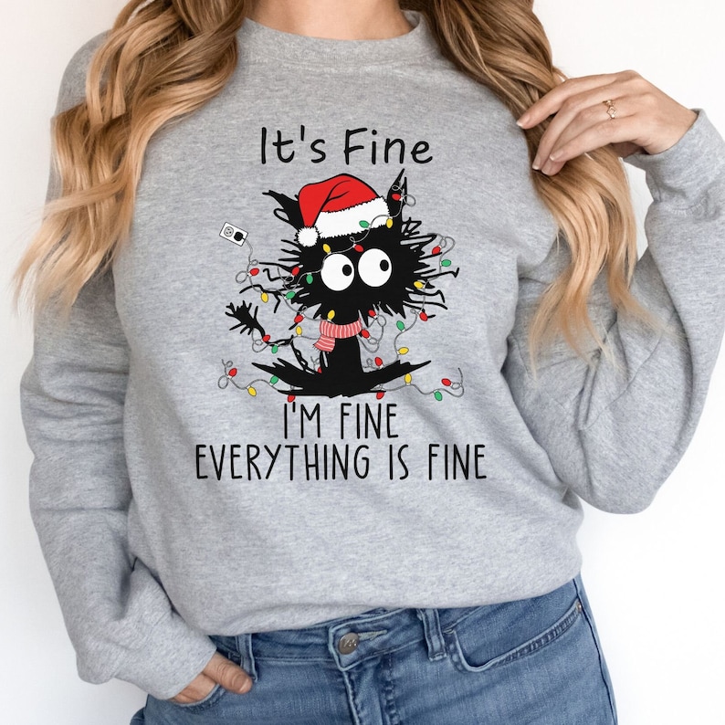 May include: A light gray sweatshirt featuring a black cat tangled in Christmas lights and wearing a Santa hat and scarf. The text "It's Fine, I'm Fine, Everything is Fine" is printed above and below the cat illustration.