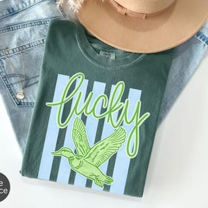 May include: A green t-shirt with the word "lucky" in a light green script, and a green duck graphic. The shirt is laid on top of blue jeans and a tan hat. A gold necklace is visible.