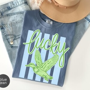 May include: A blue t-shirt with the word "Lucky" in green script and a green duck graphic. The shirt has light blue vertical stripes and is paired with light blue jeans and a tan hat.