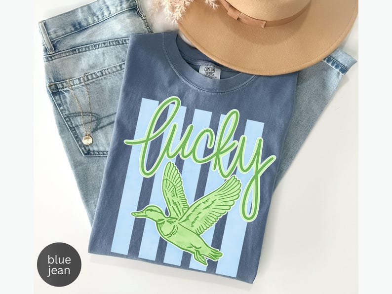 May include: A blue t-shirt with the word "Lucky" in green script and a green duck graphic. The shirt has light blue vertical stripes and is paired with light blue jeans and a tan hat.