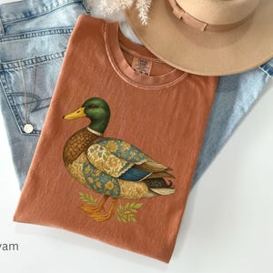 Puede incluir: Una camiseta de color terracota con una ilustraci&oacute;n detallada de un pato. El pato tiene una cabeza verde, un pico amarillo y alas estampadas. La camiseta est&aacute; colocada sobre unos vaqueros azul claro y cerca de un sombrero beige. La palabra "yam" es visible.