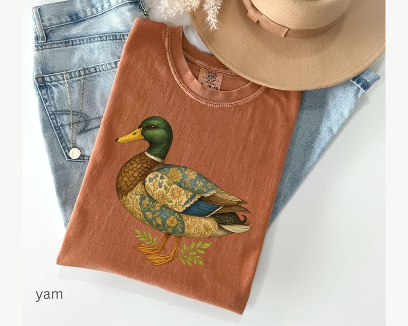 Puede incluir: Una camiseta de color terracota con una ilustraci&oacute;n detallada de un pato. El pato tiene una cabeza verde, un pico amarillo y alas estampadas. La camiseta est&aacute; colocada sobre unos vaqueros azul claro y cerca de un sombrero beige. La palabra "yam" es visible.