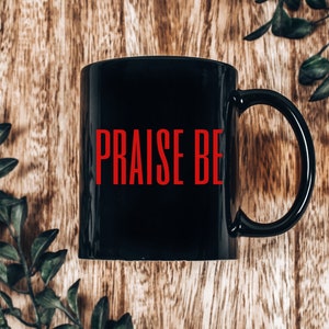 May include: Black ceramic coffee mug with a red inscription that reads "PRAISE BE". The mug is sitting on a wooden surface.
