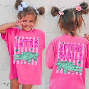 Puede incluir: Una camiseta rosa brillante con las palabras "LATER" y "AFTER WHILE" impresas en la parte delantera y trasera. La camiseta presenta un gr&aacute;fico de un caim&aacute;n verde y rayas rosas verticales. La camiseta la lleva un ni&ntilde;o peque&ntilde;o.
