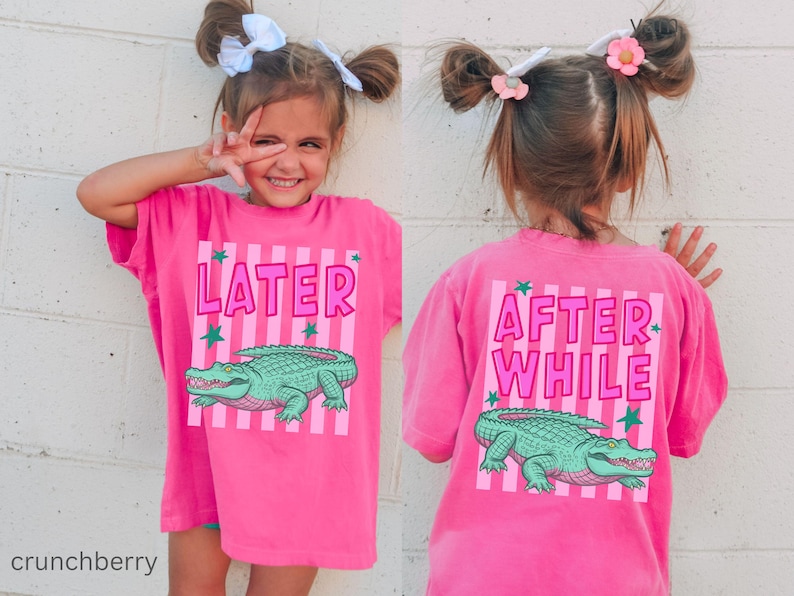 Puede incluir: Una camiseta rosa brillante con las palabras "LATER" y "AFTER WHILE" impresas en la parte delantera y trasera. La camiseta presenta un gr&aacute;fico de un caim&aacute;n verde y rayas rosas verticales. La camiseta la lleva un ni&ntilde;o peque&ntilde;o.